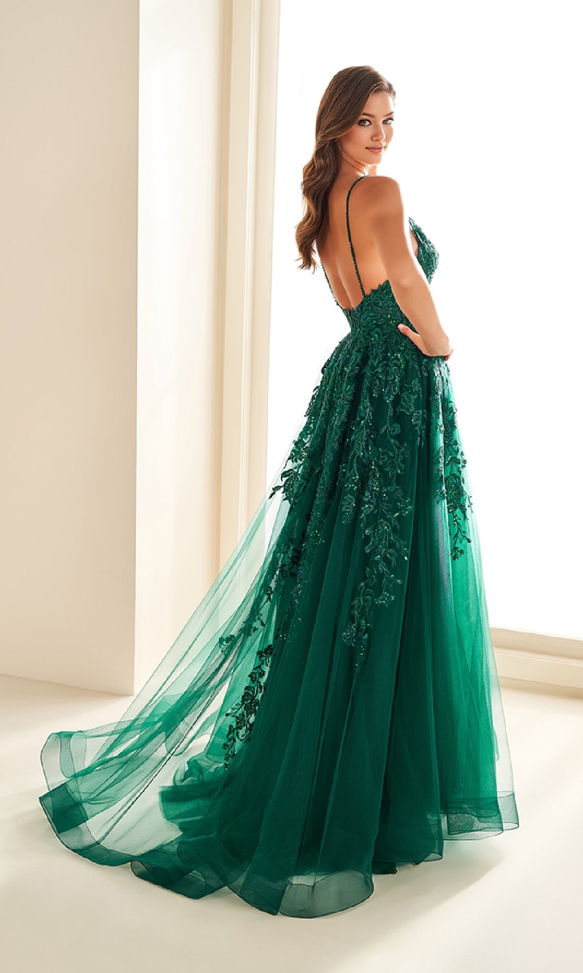 A woman stands indoors in the Ellie Wilde EW36206, a floor-length emerald green prom dress with floral embroidery, thin straps, and a corset bodice, looking over her shoulder.